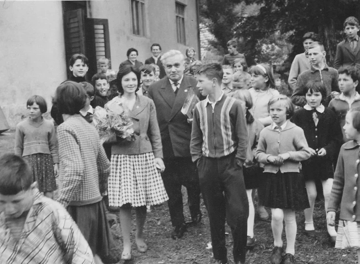 A black-and-white photograph shows a group of children gathered around two adults in front of a building. A woman is holding a bouquet of flowers, while the children stand and observe the scene, suggesting a group event or gathering.