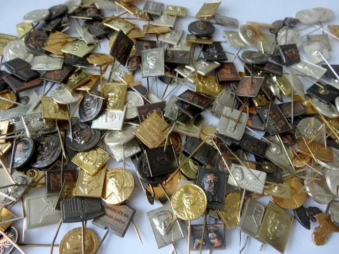 A pile of metal badges in various shapes and colors is scattered on a white surface, most featuring embossed portraits. The badges are attached to long metal pins.