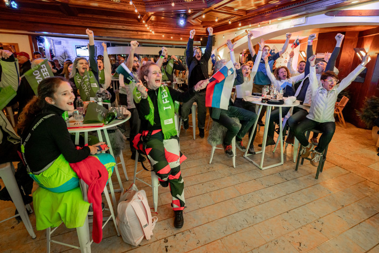 A group of enthusiastic fans in a bar cheer with raised arms. They wear colorful outfits and scarves. The atmosphere is lively and joyful.
