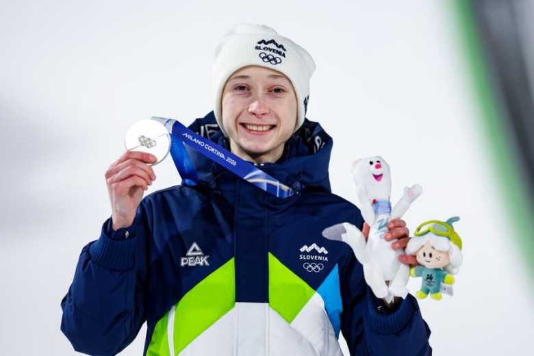 Athlete smiles while holding a silver medal and two plush toys, wearing a Slovenia jacket and beanie. The setting is a winter sports event.