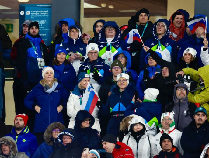 A crowd of enthusiastic individuals in blue and white winter attire cheer at a sporting event. Some hold flags and wear hats, conveying excitement and unity.