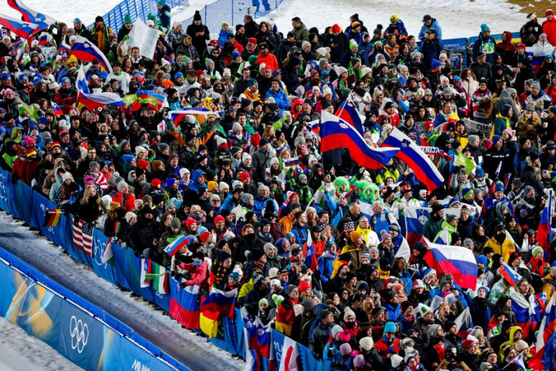 A crowd of enthusiastic individuals in blue and white winter attire cheer at a sporting event. Some hold flags and wear hats, conveying excitement and unity.