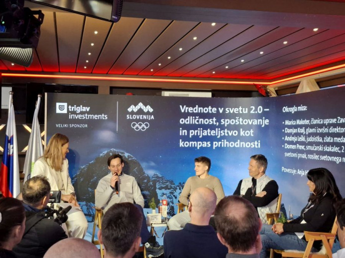 A panel of five speakers sit on stage under a sign with Slovenian text about values, sponsored by Triglav Investments. The mood is engaged and professional.