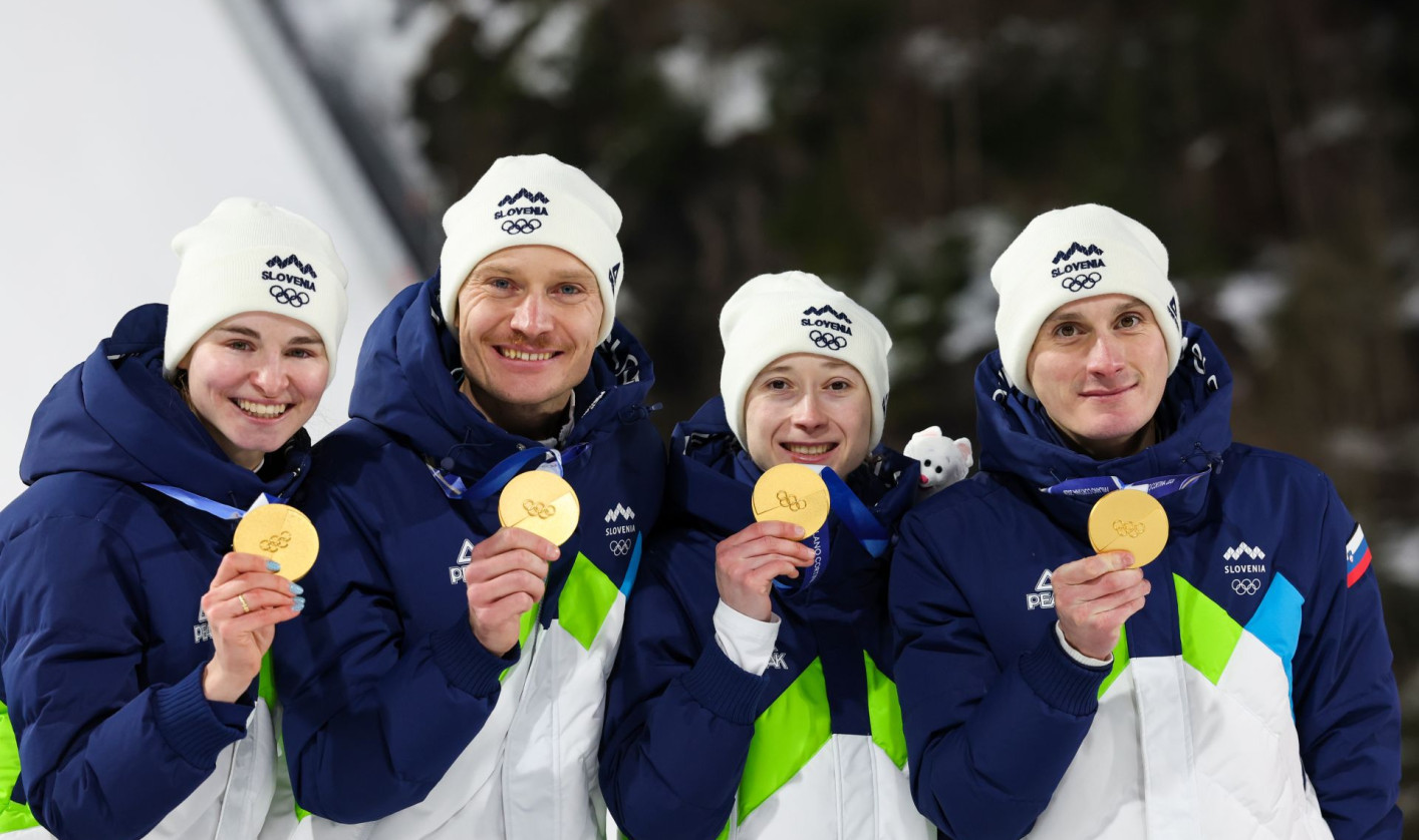 Four athletes in blue and white winter jackets proudly display gold medals, standing on a podium against a snowy backdrop, exuding joy and triumph.