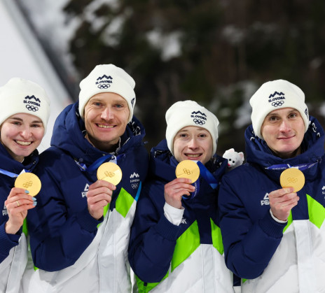 Four athletes in blue and white winter jackets proudly display gold medals, standing on a podium against a snowy backdrop, exuding joy and triumph.