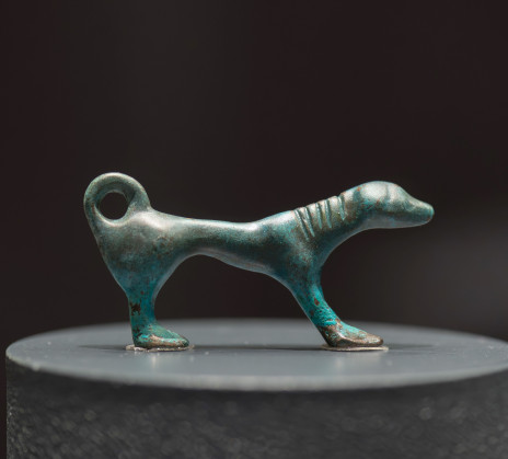 A small, turquoise patina bronze sculpture of a stylized dog with a looped tail stands on a pedestal, the background is dark.