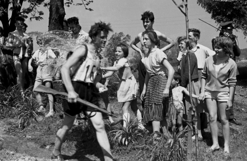 Young man holding a gun walking past a group of children and women