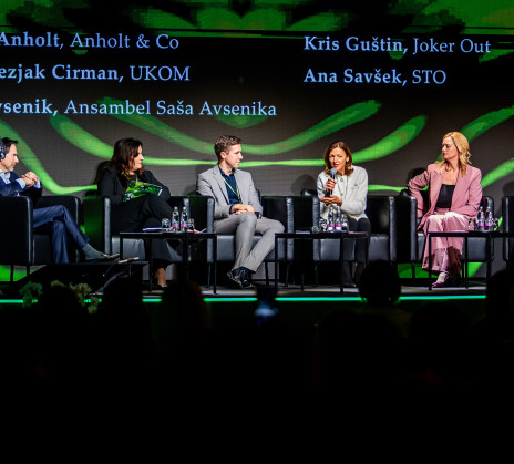 A panel discussion on stage with several participants seated in armchairs and conversing. Above them is a large screen displaying the title along with the names of speakers and the moderator.
