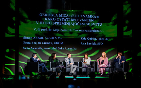 Okrogla miza na odru z več udeleženci, ki sedijo v naslanjačih in razpravljajo. Nad njimi je velik zaslon z naslovom okrogle mize in imeni govorcev in moderatorja.