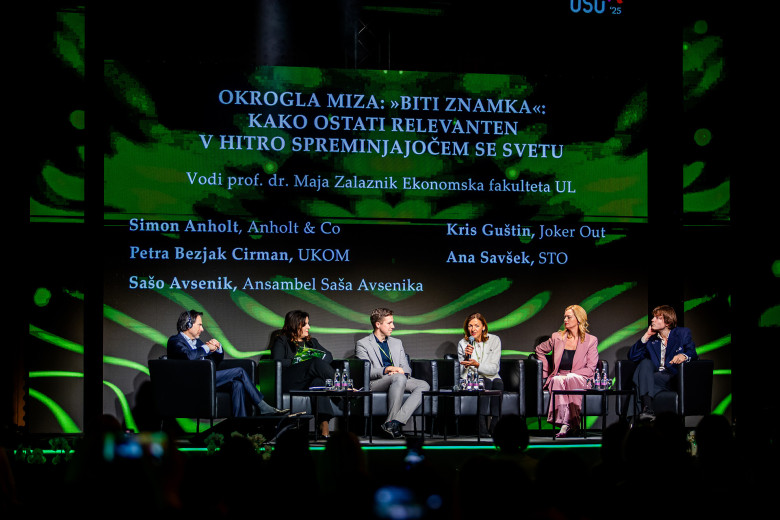 Okrogla miza na odru z več udeleženci, ki sedijo v naslanjačih in razpravljajo. Nad njimi je velik zaslon z naslovom okrogle mize in imeni govorcev in moderatorja.