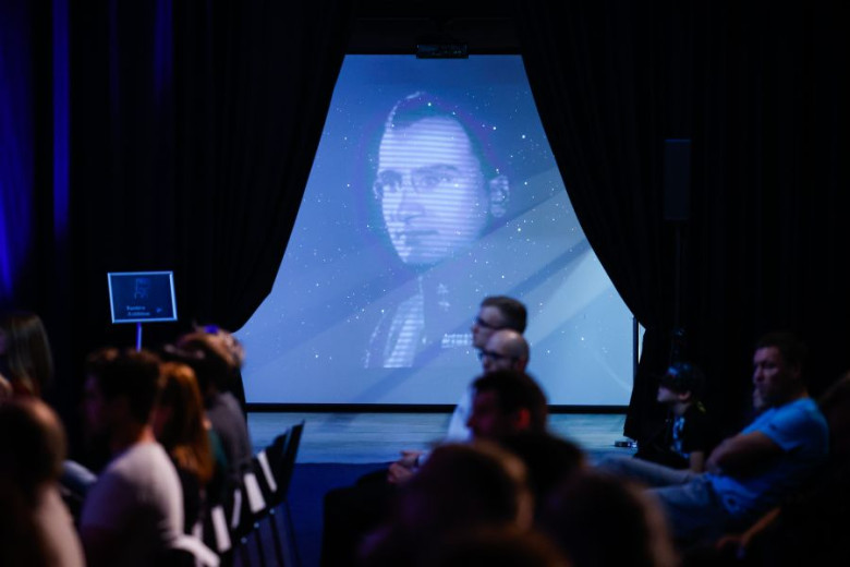 Audience seated in a darkened theater, viewing a large screen displaying an image of Herman Potočnik Noordung.