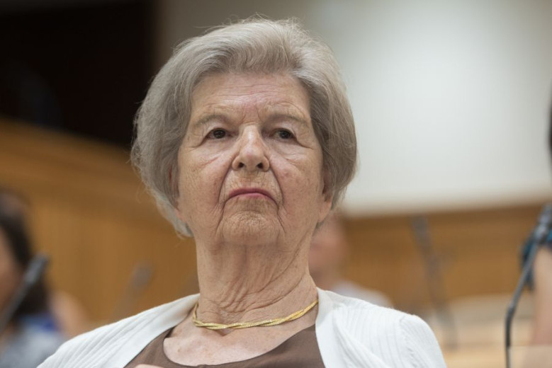 Alenka Šelih, an elderly woman with gray hair, observes attentively with a neutral expression. She wears a white cardigan.