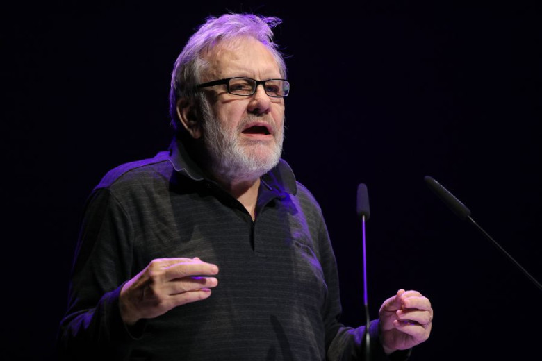 Slavoj Žižek, wearing glasses and dark clothing, speaking at a podium with microphones.
