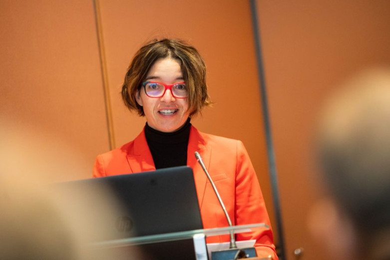 Maruša Bradač stands behind a clear podium with a microphone, wearing a red blazer, a black turtleneck, and red-framed glasses.