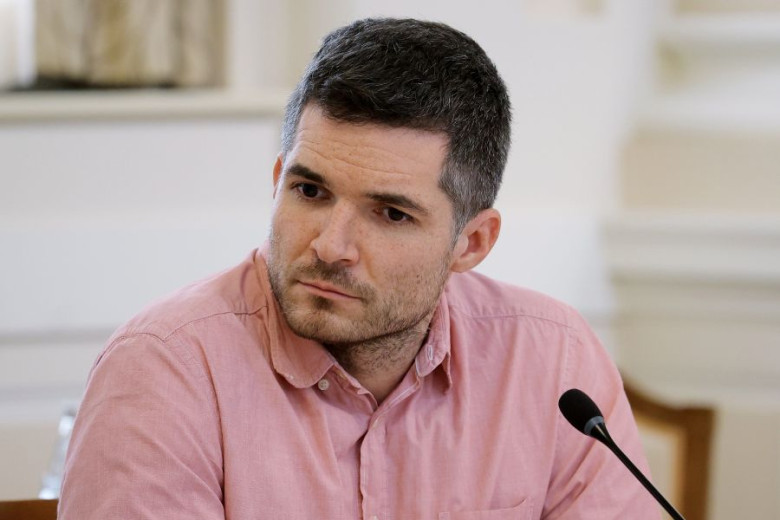 Žiga Zaplotnik, a man with dark hair and a short beard, wearing a pink button-down shirt, sits at a table with a focused expression.