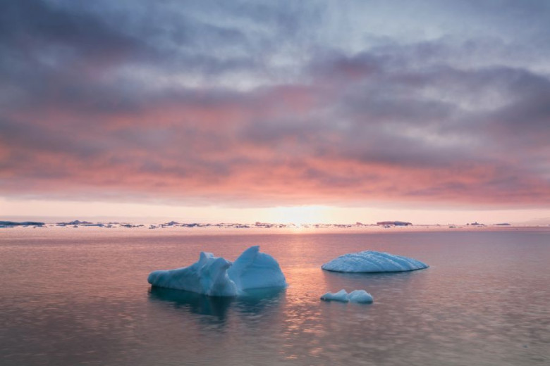 Icebergs float in calm water under a colorful sky at sunset.