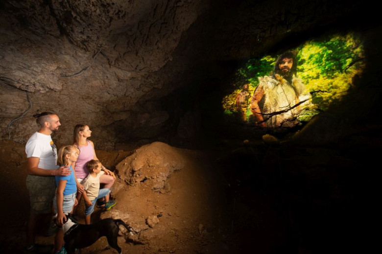 A family of four with a dog stands in a dimly lit cave, looking at a vivid projection of a prehistoric person in fur clothing against a green forest background. 