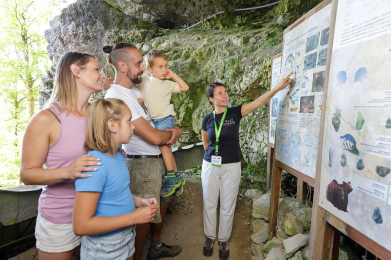 A family of four, with two children, attentively listens to a tour guide pointing at an informational board in a rocky, cave-like setting.