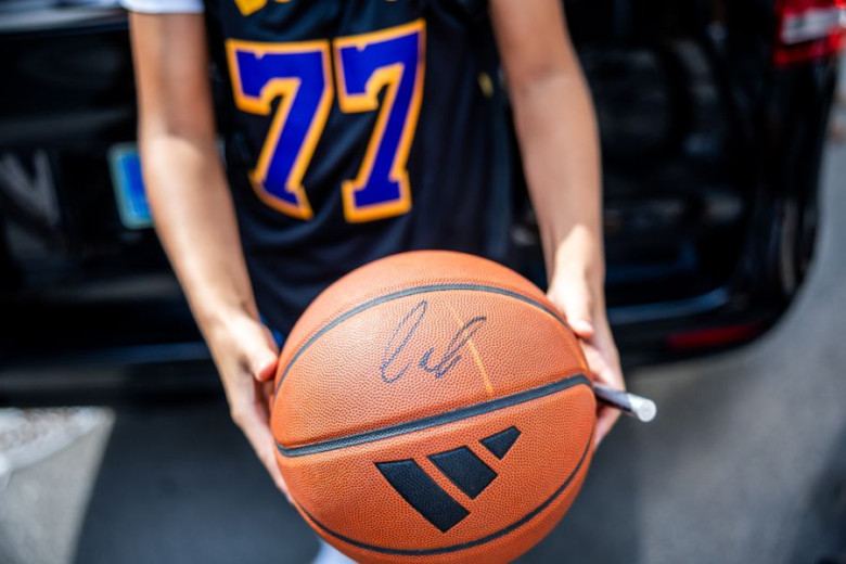 Close-up of a basketball fan in a black-and-gold number 77 jersey holding a basketball signed by Luka Dončić.