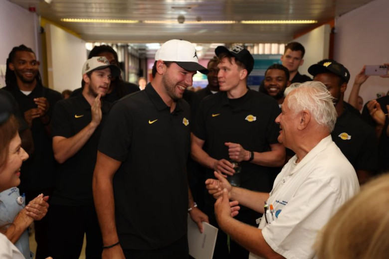 Luka Dončić smiles while talking to the obstetrician who assisted at his birth more than 27 years ago, with his teammates visible in the background.