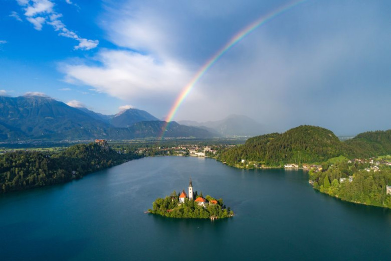 Rainbow over a blue lake Bled with a tiny church on the island in the middle with forested hills, lakeside villages, and misty mountains in the background.