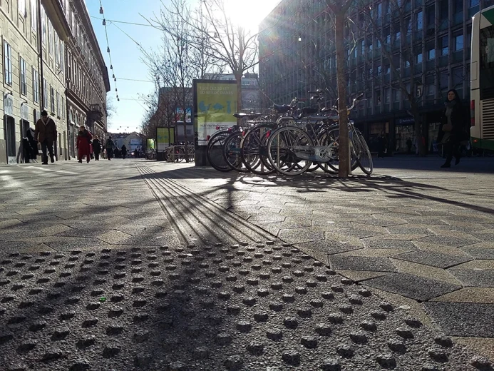 The city pavement features tactile warning surfaces and a guidance path for blind and visually impaired people. In the background are parked bicycles, pedestrians, and a city street.