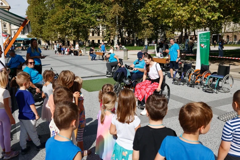 A group of children watches a demonstration of navigating obstacles in a wheelchair at a public event.