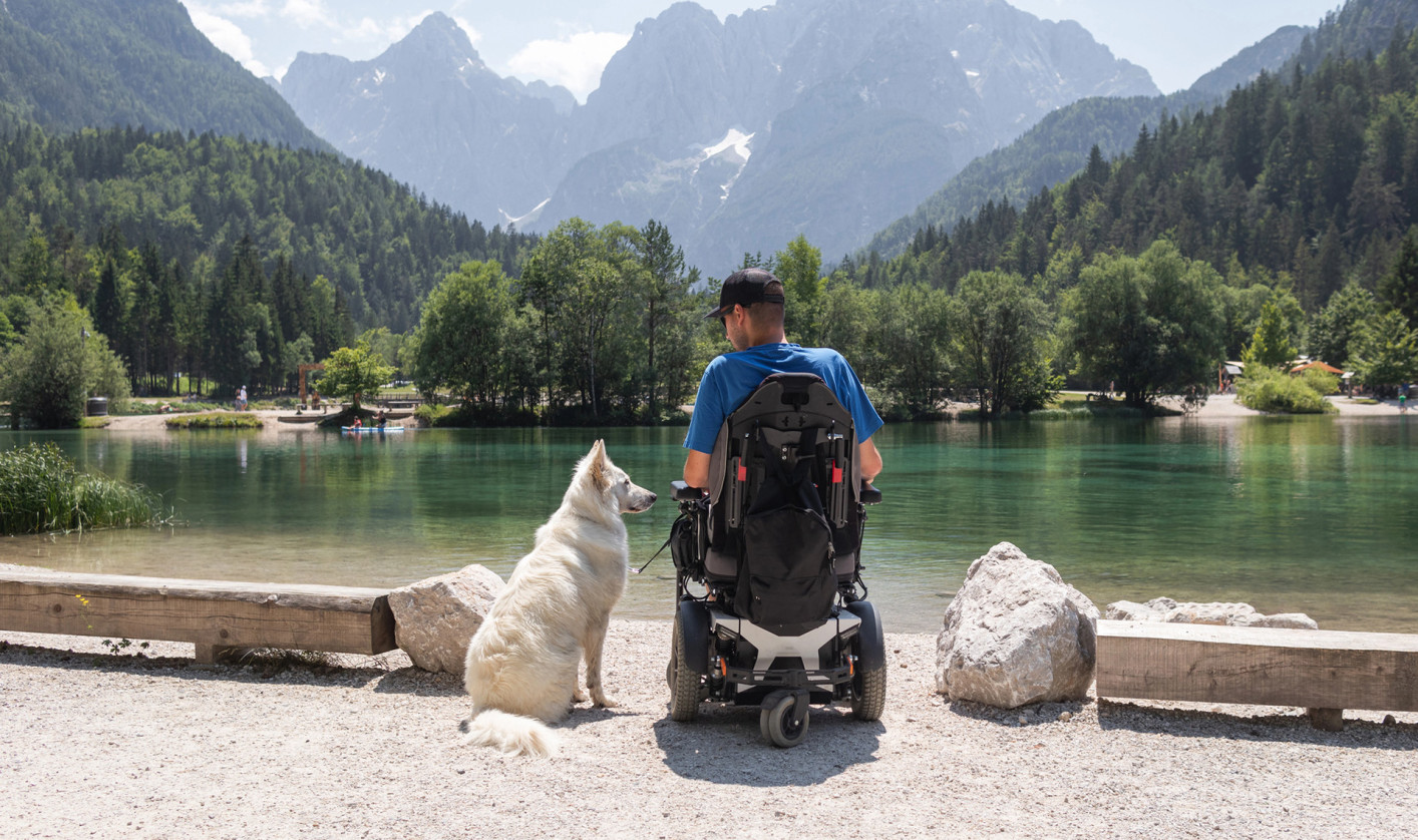 A person in a powered wheelchair sits by a lake with a dog, with the Julian Alps rising in the background.