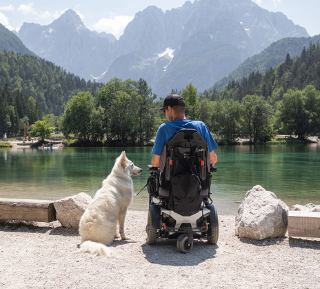A person in a powered wheelchair sits by a lake with a dog, with the Julian Alps rising in the background.