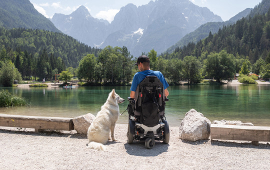 A person in a powered wheelchair sits by a lake with a dog, with the Julian Alps rising in the background.