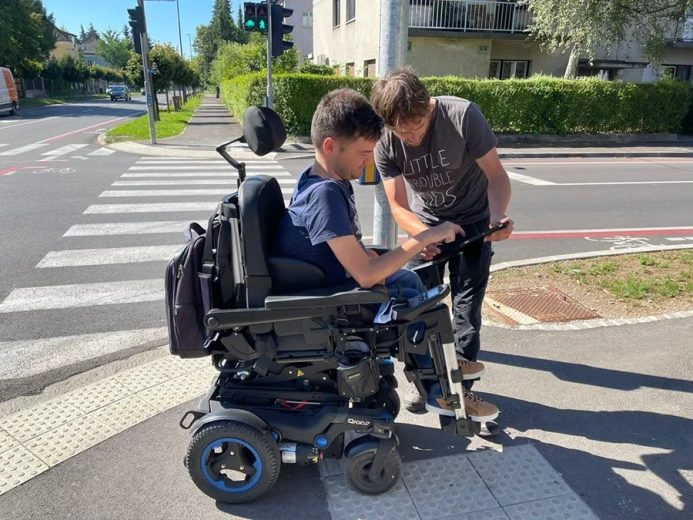 A person in a powered wheelchair and another person enter accessibility data on a tablet at a pedestrian crossing.