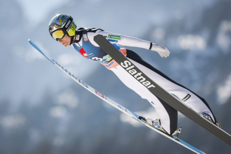 Ski jumper in mid-air, wearing white and black gear, gliding over a snowy blurred backdrop.