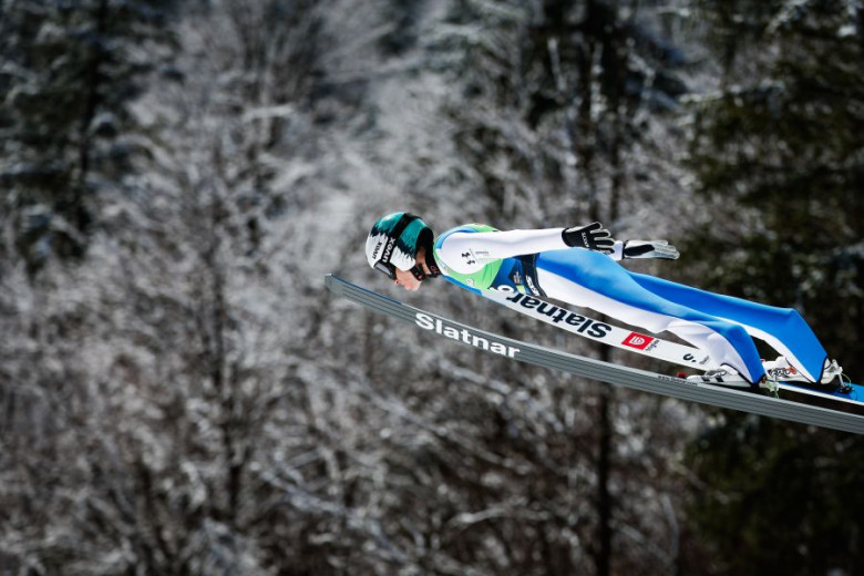 Ski jumper in mid-air, wearing a blue suit and helmet, glides over a snowy forest backdrop.