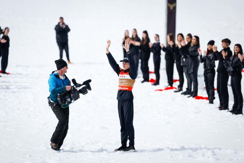 Ski jumper Peter Prevc stands with his arms raised triumphantly high in the air; in front of him is a cameraman, and behind him a line of people applauding him.