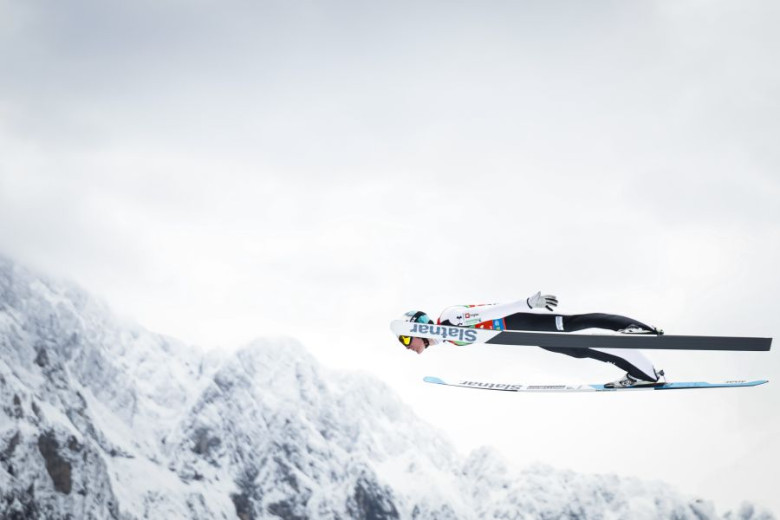 Ski jumper in mid-air, wearing white and black gear, gliding over a snowy blurred backdrop.