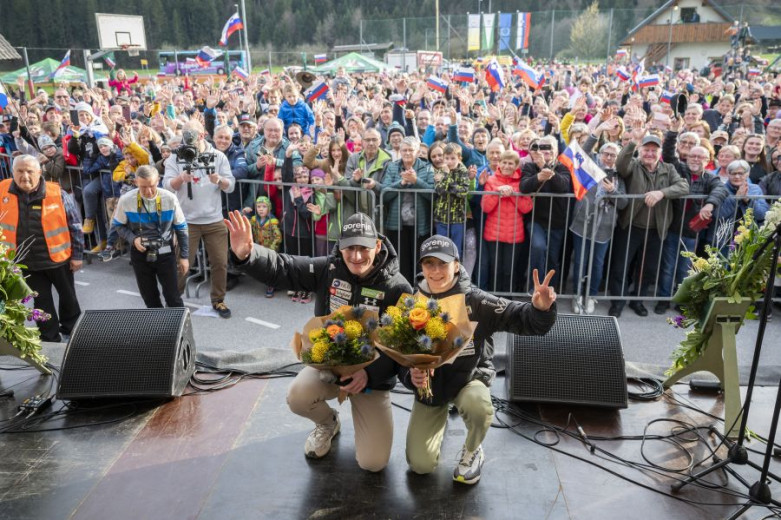 Domen and Nika Prevc on stage holding flowers, smiling. A large, cheering crowd waves flags behind them.