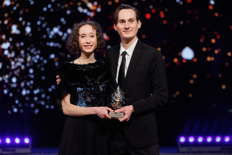 Smiling Nika and Domen Prevc, dressed in formal black outfits, are holding a small trophy in their hands.