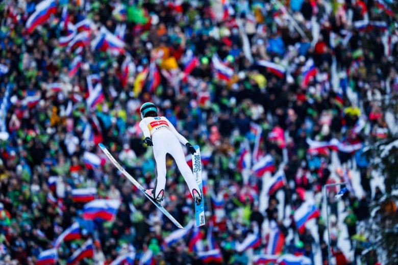 A ski jumper in mid-air wearing a white suit and helmet, with a large crowd in the background.