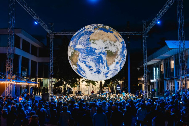A large illuminated globe suspended above a crowd at an outdoor night-time festival, with blue stage lighting and surrounding buildings.