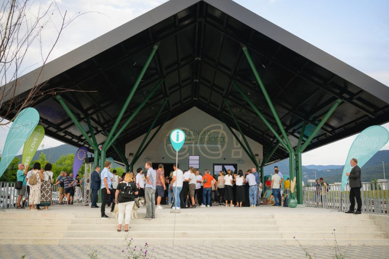 A group of people is standing outdoors under a roof supported by green pillars.