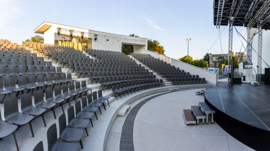 Empty outdoor amphitheater with tiered seating and stage, bathed in sunlight.