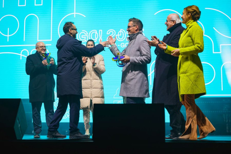 Event attendees applaud as ttwo man skae hands on stage. 