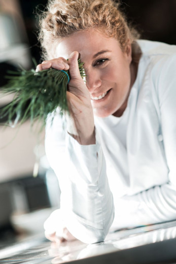 A smiling Ana Roš holding herbs and hiding her eye under them.