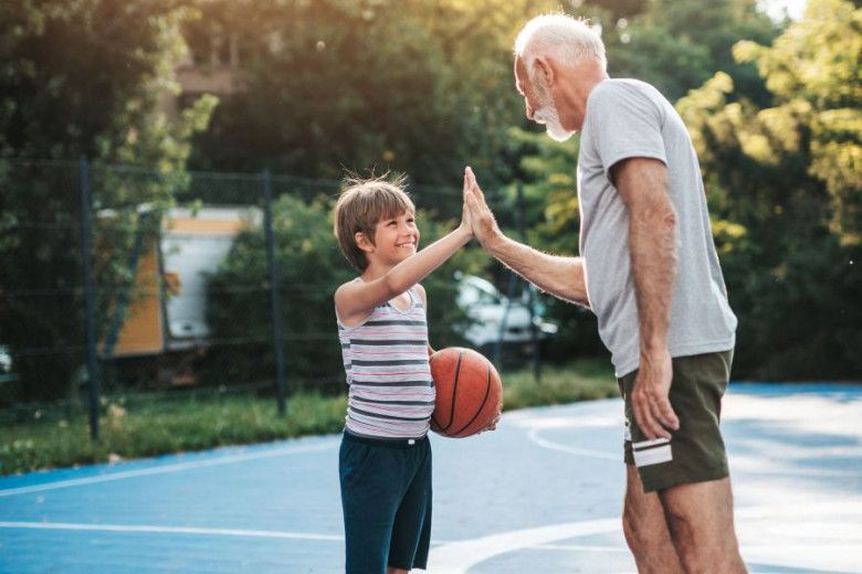A grandson with a basketball and a grandfather on the court give each other a high five.