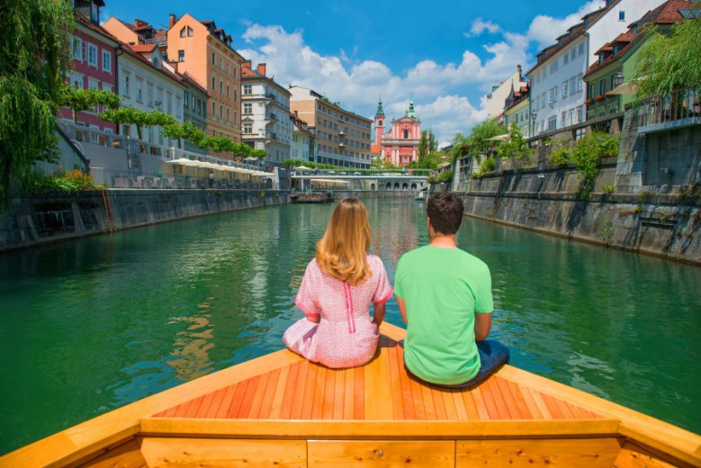 A couple is sitting on a boat on the river. They look towards the town houses and the church.
