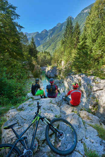 Trije kolesarji počivajo na skali s pogledom na turkizno reko.