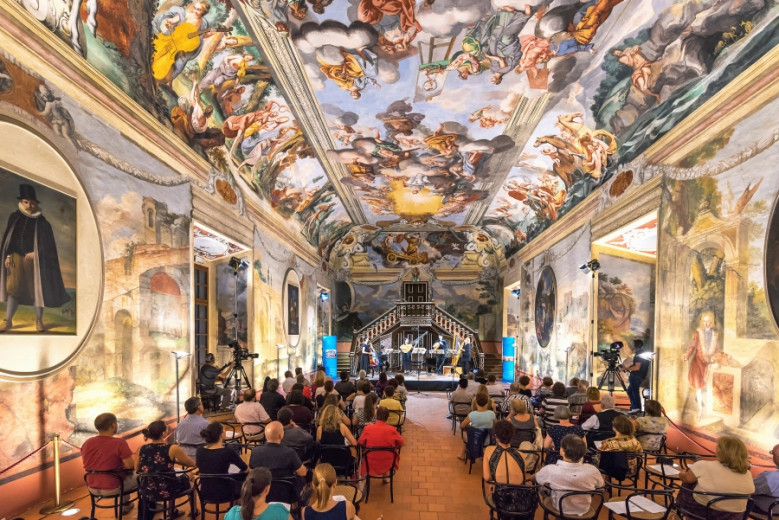 People sitting in a frescoed hall and watching a musical performance.
