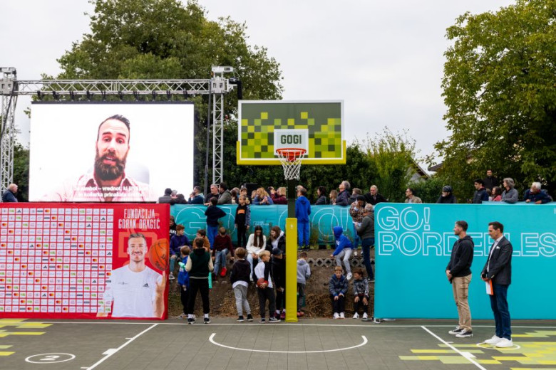 A man on a television screen is addressing the crowd by the basketball court.