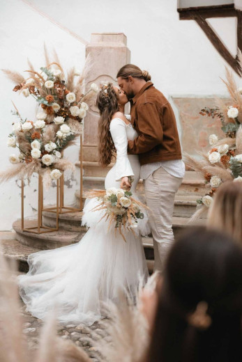 A bride and groom kiss on stone stairs at an outdoor wedding. The bride wears a white dress with long sleeves and a flower crown. The groom wears a brown jacket and light pants. Floral displays of pampas grass and white roses frame the couple.