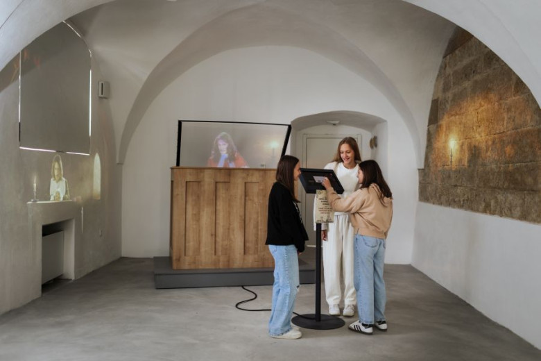 Three visitors are looking at a screen with instructions for participating in a trial. Behind them is a wooden desk, and two judges are projected on the walls. The room is bright, with white walls and an arched ceiling.
